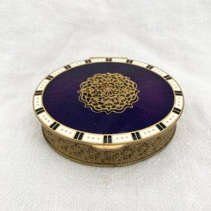 May include: A round, gold-coloured compact with a purple enamel top. The top features a decorative, gold-coloured design in the centre and a black and white border.