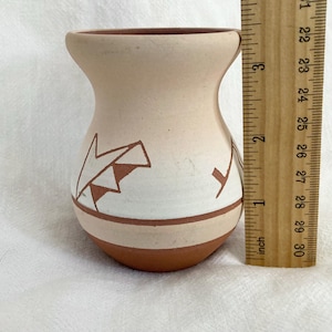 May include: A small, beige ceramic vase with a rounded base and a flared top. The vase features geometric designs in brown and white. The vase is approximately 7.6 cm tall.