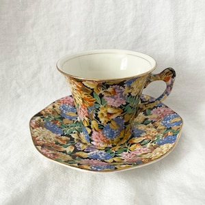 以下が含まれることがあります： 黒い背景に、黄色、ピンク、青、オレンジの色合いの花柄が施された、ヴィンテージのティーカップとソーサーのセット。カップとソーサーには金のトリムが付いています。