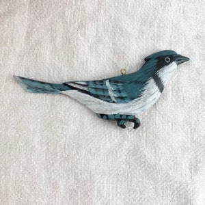 May include: A hand-painted wooden blue jay bird ornament with white accents. The bird is perched on a branch with its wings spread out.
