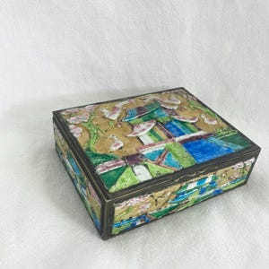 May include: A rectangular metal box featuring a raised, colorful enamel scene on the lid and sides. The design depicts a landscape with a pagoda, green trees, pink blossoms, and white birds against a tan background, finished with a dark metal trim.