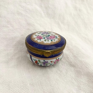 May include: A small, round porcelain trinket box with a blue and gold lid. The lid is decorated with a floral design in pink, white, and purple. The box is approximately 5 cm in diameter.