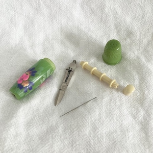 May include: A selection of sewing tools, including a green floral needle case, small scissors, a thimble, a needle, and a bone-coloured pin cushion. The items are arranged on a white, textured surface.
