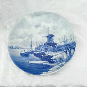 May include: A round, blue and white decorative plate featuring a winter landscape scene. The image depicts a windmill, boats, and buildings along a body of water. The plate has a glossy finish and a detailed, hand-painted design.