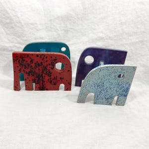 Puede incluir: Cuatro figuritas de elefante de cerámica en diferentes colores. Los elefantes están de pie en fila, con el elefante rojo en la parte delantera, seguido por el azul, el morado y el azul claro.