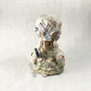 May include: A ceramic mouse figurine wearing a ruffled bonnet with blue and pink accents. The mouse has a brown face and holds a small grey circular object. The dress features a cream base with colorful floral patterns and ruffled edges.