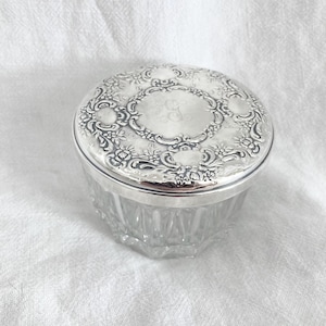 May include: A vintage silver-plated vanity jar with a clear, faceted glass base. The lid is intricately decorated with floral and scroll patterns, and features a central monogram. The jar is round and sits on a white surface.