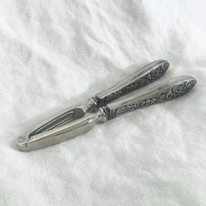 May include: A silver nutcracker with ornate floral and scrollwork detailing on the handles. The nutcracker is open, revealing the serrated jaws designed to crack nuts. The handles are engraved with the text "Reed & Barton America".