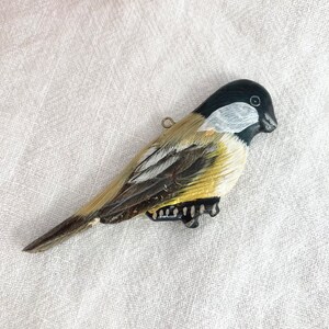 May include: A hand-painted wooden bird ornament with black, white, and yellow details. The bird has a black head with a white stripe and a yellow body with black markings.