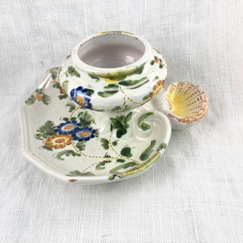 May include: A white ceramic candle holder with a floral design. The holder has a round base with a small dish on the side. The holder is decorated with yellow, blue, and green flowers.