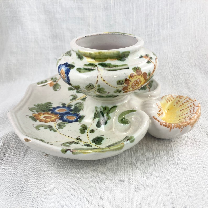 May include: A white ceramic candle holder with a floral design. The holder has a central cup and two smaller side cups. The floral design is in shades of blue, yellow, and green.