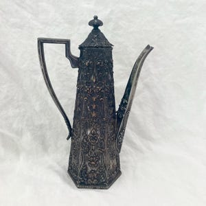 Puede incluir: Una cafetera antigua de color plateado con intrincados detalles florales y de volutas. La cafetera presenta un cuerpo alto y hexagonal, un pico curvo y un asa. La tapa está rematada con un adorno decorativo.