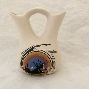 May include: A white ceramic Native American style vase with a black and brown geometric design. The vase has a handle and two spouts.
