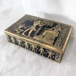 May include: A rectangular box featuring a black surface with gold-toned Egyptian-style engravings. The lid displays figures in profile and hieroglyphics, while the sides are decorated with a continuous frieze of standing human figures.