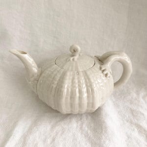 May include: Off-white ceramic teapot with a ribbed, pumpkin-like body. The teapot has a curved spout, a loop handle, and a lid with a small, decorative knob. The teapot is sitting on a white, textured surface.