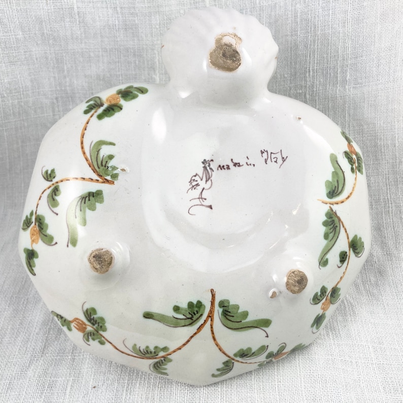 May include: White ceramic dish with green and gold floral designs. The bottom of the dish has three feet and a handwritten inscription that reads "ma fa ti, Italy".