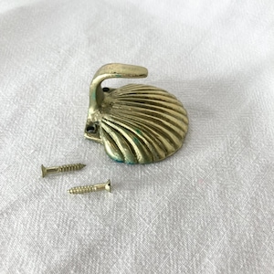 May include: A brass-toned metal wall hook shaped like a seashell with a curved hook extending from the top. The shell base features a ribbed texture and two small holes for mounting. Two matching metal screws are positioned next to the hook.