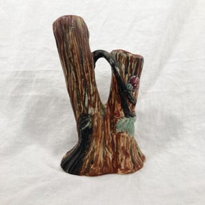 May include: A ceramic vase shaped like a tree trunk with two branches. The vase is painted in shades of brown, green, and red to resemble bark and leaves.