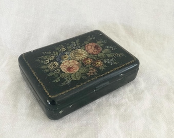 May include: A small black box with a floral design painted on the lid. The flowers are pink, yellow, and white, and the box has a gold trim.