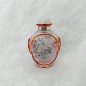 Puede incluir: Una pequeña botella de vidrio transparente con un diseño floral. La botella tiene una forma redondeada con un borde y detalles en rojo. El diseño floral presenta flores rosas y hojas verdes. La botella está rematada con un tapón blanco.