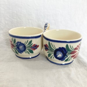 Könnte beinhalten: Cremefarbenes Keramik-Serviergeschirr mit zwei verbundenen Schalen und einem Griff. Jede Schale ist mit einem blauen Blumendesign, grünen Blättern und roten Akzenten verziert. Die Ränder sind mit einem blauen Band versehen. Ein dekoratives Stück zum Servieren.