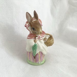 May include: A ceramic figurine of a rabbit dressed in a white and pink dress, holding a green umbrella and a woven basket. The rabbit has brown fur and is wearing a red and white bonnet. The figurine is approximately 4 inches tall.