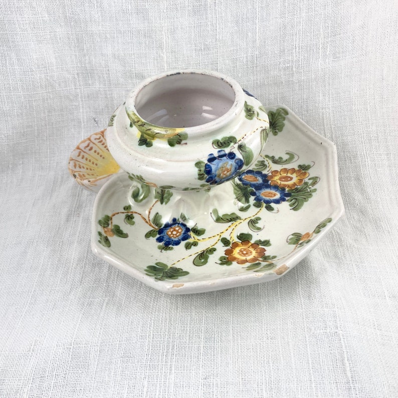 May include: A white ceramic dish with a floral design in shades of blue, yellow, and green. The dish has a scalloped edge and is decorated with a floral pattern. The dish is sitting on a white fabric background.