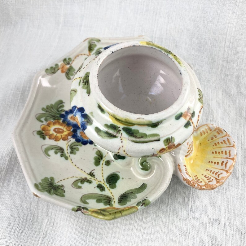 May include: A white ceramic dish with a floral design in shades of blue, yellow, and green. The dish has a raised edge and a scalloped handle. The dish is decorated with a floral pattern in shades of blue, yellow, and green.