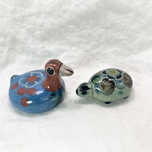 May include: Two ceramic ornaments: a duck and a turtle. The duck is blue with orange and white floral accents, a brown head, and a gray beak. The turtle is light green with floral patterns and black accents.
