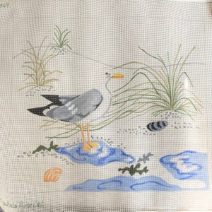 May include: A needlepoint canvas with a gray and white seagull standing on a blue and white water design. The seagull is surrounded by green grass and a seashell.