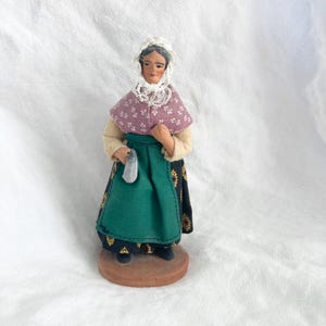 May include: A vintage figurine of a woman in traditional attire. She wears a white bonnet, a pink cape with white floral patterns, and a green apron over a black skirt with gold accents. The figurine is mounted on a brown base.