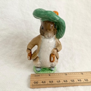 May include: A ceramic figurine of Peter Rabbit wearing a green hat and brown jacket, holding a carrot in each hand. The figurine is approximately 7.5 cm tall.