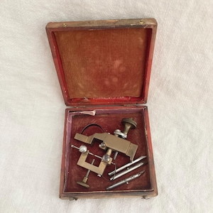 May include: Antique brass tool in a wooden box with a red velvet lining. The tool has a handle, a clamp, and three metal rods.