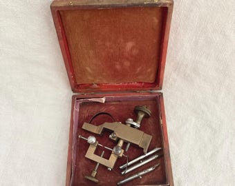 May include: Antique brass tool in a wooden box with a red velvet lining. The tool has a handle, a clamp, and three metal rods.