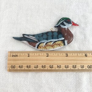 May include: Hand-painted wooden ornament of a wood duck with a green head, red beak, and brown, blue, and gold feathers. The ornament is approximately 2 inches long and is measured against a ruler.