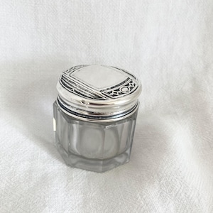 May include: A small, antique glass jar with a silver lid. The jar is octagonal and clear, while the lid is silver with an ornate geometric design. The lid has a small, circular hole. The jar sits on a white surface.
