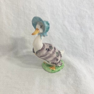 May include: A white ceramic figurine of a duck wearing a blue hat and a purple and green patterned coat. The duck is standing on a green base.