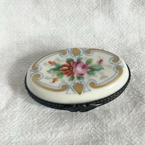 Könnte beinhalten: Ovale Schmuckdose aus Porzellan mit floralem Design. Der Deckel zeigt eine zentrale Anordnung aus rosa und orangefarbenen Rosen mit grünen Blättern, umrahmt von goldenen Akzenten und einem blauen Rand. Die Dose hat einen dunklen Metallsockel.