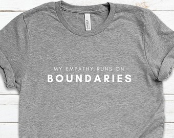 May include: A heather gray t-shirt with rolled-up sleeves and a knotted side. The shirt features the white text "MY EMPATHY RUNS ON BOUNDARIES" in a simple, sans-serif font. The shirt is made of a soft material.