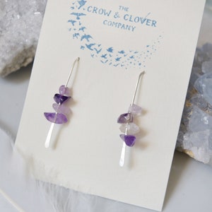 May include: A pair of silver earrings with amethyst stones. The earrings are displayed on a white card with the text "The Crow & Clover Company".