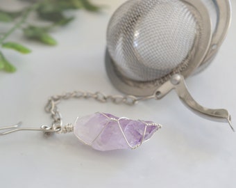 May include: A silver tea infuser with a purple amethyst crystal attached to a chain. The infuser has a round mesh ball and a handle.