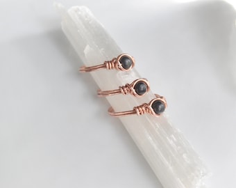 May include: Three simple rings with black gemstone beads wrapped in rose gold wire. The rings are displayed on a white crystal.