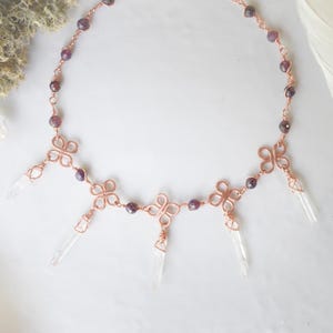 May include: A copper-colored necklace with a series of four-leaf clover-shaped designs, connected by a chain of small, round, dark red beads. Clear crystal points hang from the clover shapes. The necklace is displayed on a white surface.