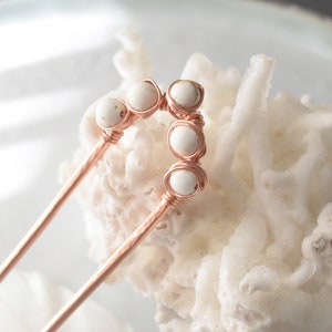 May include: A pair of rose gold hair pins with white stone accents. The stones are arranged in a curved shape on the top of each pin.