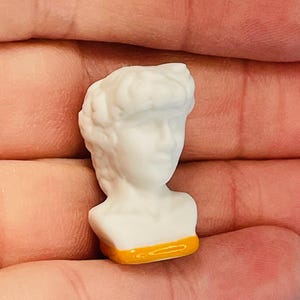May include: A small, white bust of a classical figure with detailed facial features and curly hair. The bust has an orange band at the base. The miniature sculpture is held in a hand, suggesting its small size. The bust is likely a decorative item.