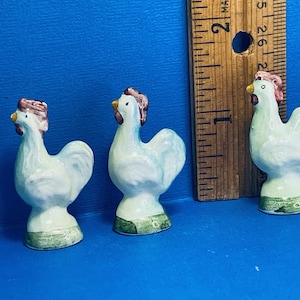 May include: Three ceramic rooster figurines. The roosters are white with blue accents, a red comb, and a yellow beak. They stand on a green base. A wooden ruler is next to the figurines. The background is a solid blue.
