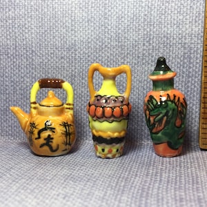 Puede incluir: Tres figuritas de cerámica en miniatura: una tetera amarilla y naranja con caracteres chinos negros, una ánfora amarilla y naranja con detalles negros y rojos, y un jarrón verde y naranja con un diseño de dragón.