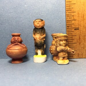 May include: Three small ceramic figurines, each depicting a different character. The first is a brown pot with a face, the second is a brown and black figure with a face, and the third is a brown figure with a face and a hat, holding a round object.