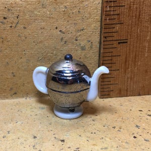 May include: A small, silver teapot with a white base and white handle. The teapot is about 2 inches tall.