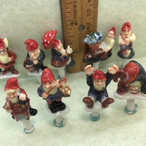 May include: Set of ten ceramic gnome figurines, each with a different pose and holding a different object. The gnomes are painted in various colours, including red, blue, green, and yellow. They are all standing on small white bases.
